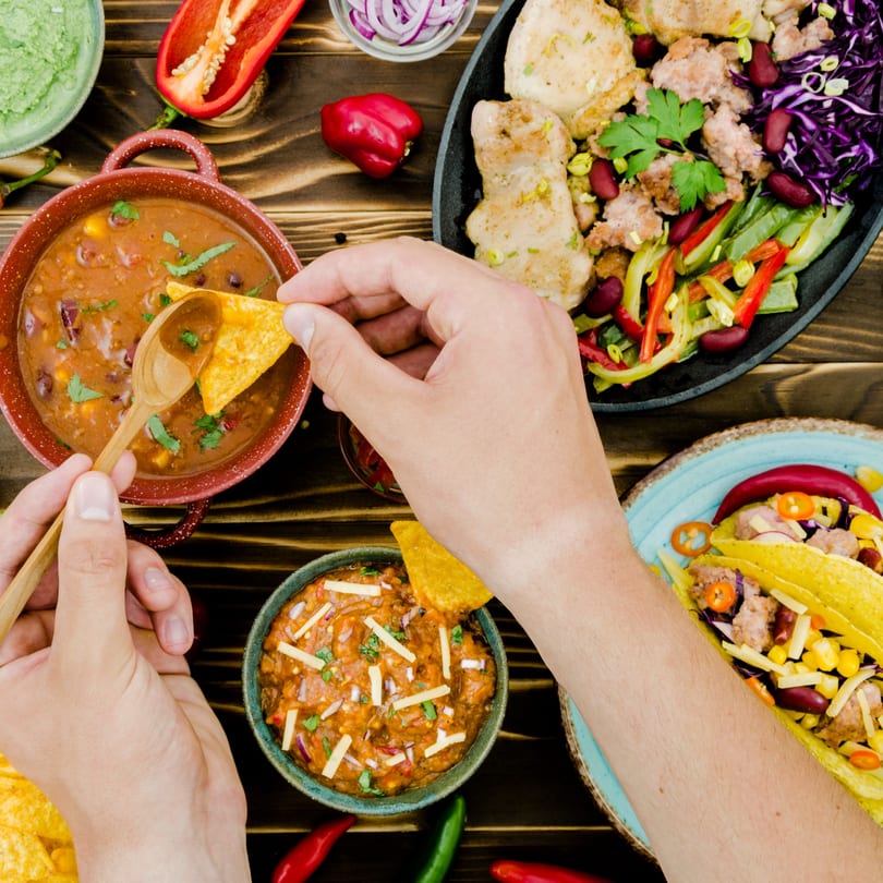 Overhead view of hands dipping tortilla chips into bowls of chili and guacamole, with tacos and fajitas.