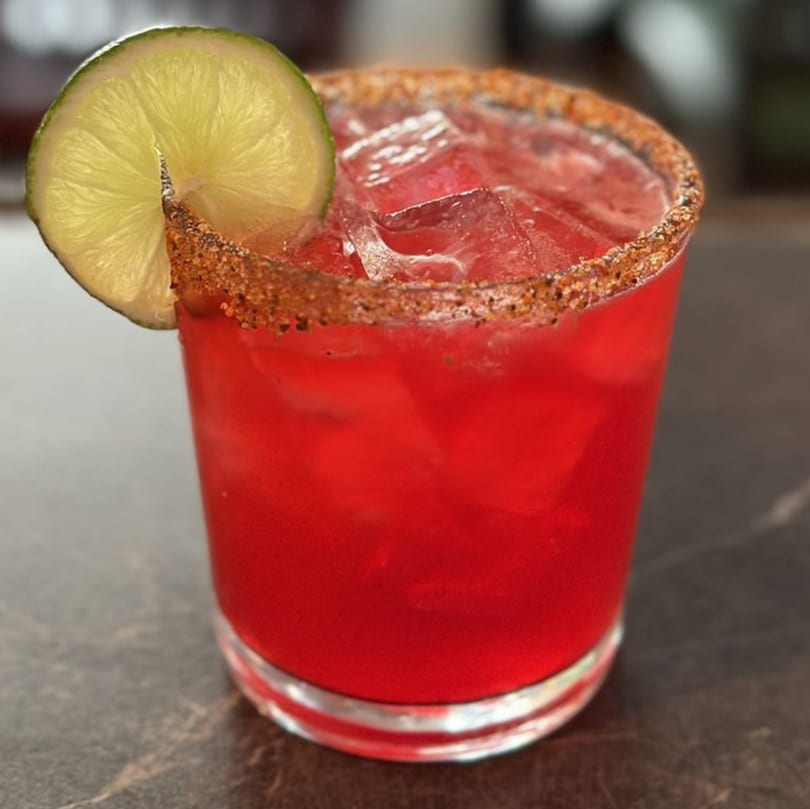 A vibrant red Mexican cocktail with ice, a lime wedge, and a spiced rim.