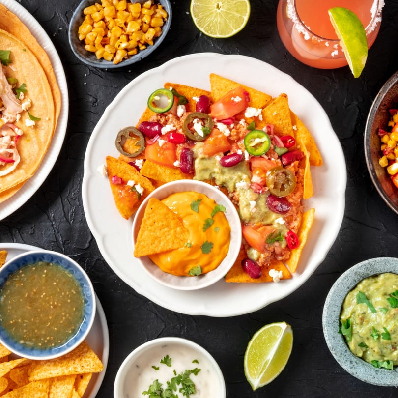 Overhead view of Maestros’s Mexican grill's nachos, tacos, dips, and drinks on a dark table.