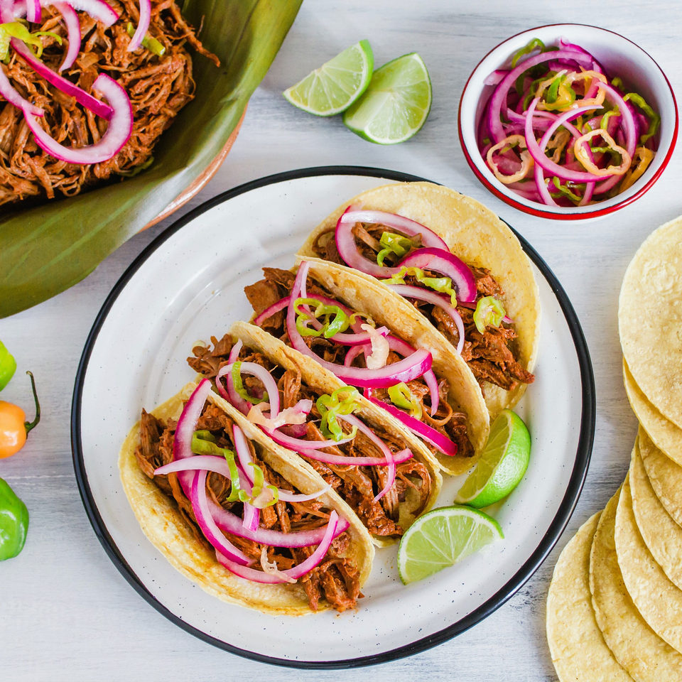 Three Maestros’s Mexican grill tacos with shredded meat, red onions, and peppers, with limes and tortillas.