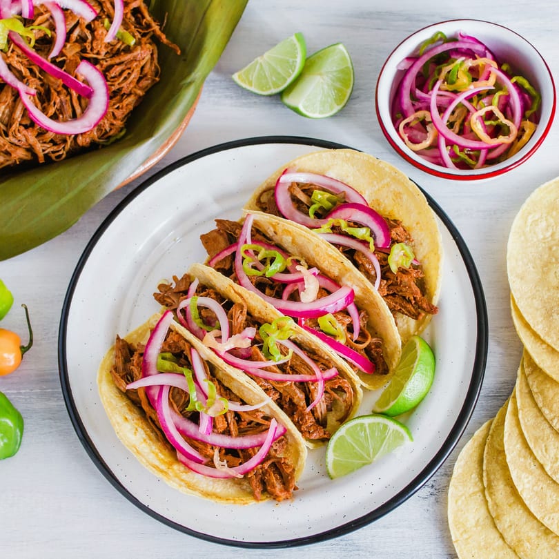 Three Maestros’s Mexican grill tacos with shredded meat, red onions, and peppers, with limes and tortillas.